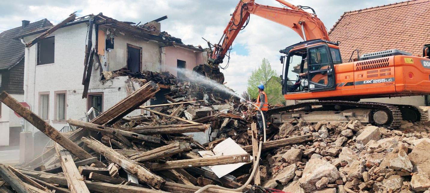 Mitarbeiter sprüht Wasser auf einem halb abgerissenen Hauses und Bagger  räumt den Schutt zur Seite